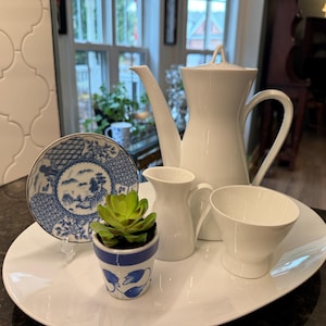 May include: A white porcelain coffee pot, creamer, and cup set on a white oval platter. A blue and white decorative plate and a small potted succulent are also on the platter. The set is displayed on a dark countertop.