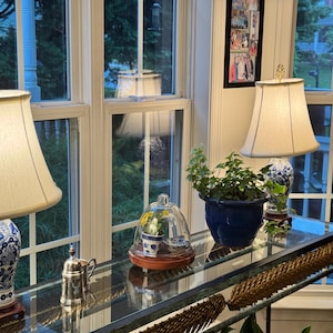 May include: A glass-topped console table displays decorative items. Two table lamps with cream-colored shades and blue and white patterned bases flank the table. A cloche, a silver creamer, and a blue planter with greenery are also visible.