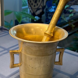 May include: A gold-toned metal mortar and pestle set featuring a wide, flared rim, two square-shaped side handles, and horizontal engraved line details around the cylindrical body. The matching pestle rests inside the basin of the mortar.