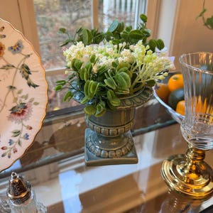 May include: A small, sage green urn-shaped vase filled with faux white and green flowers and foliage. The vase sits on a glass surface, next to a decorative plate and a glass with a gold base. The scene is set near a window.
