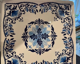 May include: Square ceramic serving platter with a cream-colored background and a blue floral design. The platter features a central floral medallion and a border of stylized leaves and flowers in varying shades of blue.