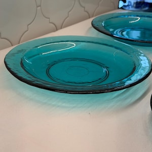 Pode incluir: Dois pratos redondos de vidro translúcido em tom azul-esverdeado com um centro plano e liso e uma borda elevada ligeiramente texturizada. O vidro tem um tom azul-esverdeado profundo e um acabamento brilhante que reflete a luz na superfície.