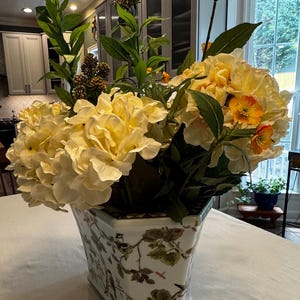 May include: A floral arrangement in a white and patterned ceramic vase. The arrangement features large, creamy yellow flowers, green leaves, and accents of orange blooms. The vase has a rectangular shape with a detailed botanical design.