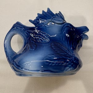 Könnte beinhalten: Eine kobaltblaue Keramik-Sahnekännchen mit einem dekorativen Drachendesign. Das Sahnekännchen hat einen Griff und einen Ausguss, wobei der Drachenkopf die Oberseite bildet. Ein einzigartiges Stück zum Servieren von Sahne oder anderen Flüssigkeiten.