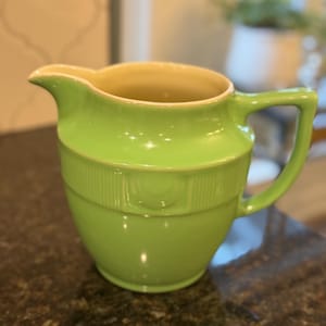 May include: A bright green ceramic creamer with a handle and spout. The creamer has a ribbed band around the middle and a light yellow interior. The creamer is sitting on a dark brown surface.