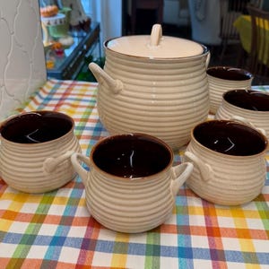 May include: A ceramic set consisting of one large lidded pot and four matching bowls, all featuring a cream-colored exterior with horizontal ribbed textures and dark brown interiors. Each piece includes small side handles for easy gripping.