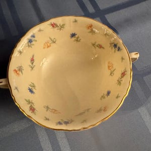 Könnte beinhalten: Cremefarbene Suppenschüssel mit einem Blumenmuster in Blau, Orange und Rosa. Der Rand ist gewellt und mit Gold verziert. Zwei kleine Griffe befinden sich auf beiden Seiten der Schüssel.