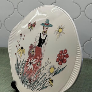 May include: A white ceramic plate featuring a colorful illustration of a person in a hat, black bodice, and red skirt standing among stylized flowers, a butterfly, and a sun. The plate has a ribbed edge and a glossy finish on the surface.