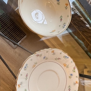 May include: Cream-colored teacup and saucer set with a floral pattern. The cup has a handle and a gold rim, and the saucer has a scalloped edge. The floral design includes blue, yellow, and orange flowers with green leaves. A classic tea set.