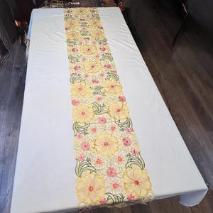 May include: A white tablecloth with a floral table runner. The runner features a repeating pattern of large yellow flowers with pink centers and green leaves. The flowers are embroidered.