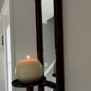 May include: A wall-mounted candle holder with a rectangular mirror. The dark brown frame has a bamboo-like design. A large, round, cream-colored candle sits on a black metal tray, with a lit flame.