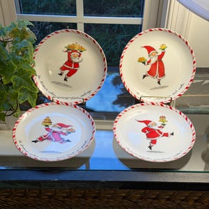 Puede incluir: Un juego de cuatro platos de cerámica blanca con un borde a rayas rojas y blancas tipo bastón de caramelo. Cada plato presenta una ilustración central de Santa Claus sosteniendo postres, acompañada de texto en cursiva en el borde.