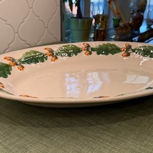 Pode incluir: Uma travessa de servir oval em cerâmica com uma base de cor creme e uma borda decorativa com folhas de carvalho verdes e pequenas bolotas cor de laranja. A travessa tem um acabamento liso e brilhante e uma base larga e rasa.