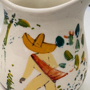 May include: White ceramic jug with a hand-painted design of a figure in a yellow outfit and sombrero, with a red sash. The jug is decorated with colourful foliage and is likely a decorative item.