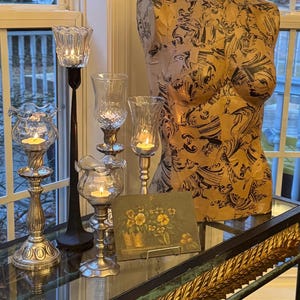 Könnte beinhalten: Eine künstlerische Inszenierung mit einer dekorativen Büste mit Muster, umgeben von beleuchteten Kerzenhaltern verschiedener Stile. Ein kleines Blumenbild und ein goldverzierter Tisch tragen zum eleganten Ambiente bei.
