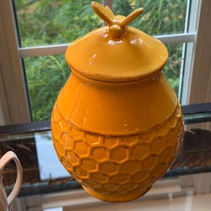 May include: A bright yellow ceramic honey jar with a rounded body and a matching lid. The exterior features a raised hexagonal honeycomb pattern across the lower section. A small, sculpted bee figure sits atop the center of the lid.