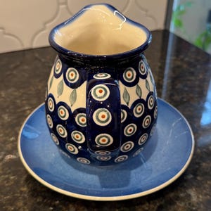 Könnte beinhalten: Eine Keramikkrug mit passender Untertasse. Der Krug ist dunkelblau mit weißem Innenraum und Henkel. Die Außenseite ist mit einem sich wiederholenden Muster aus blauen und roten Kreisen und grünen Blattformen verziert. Die Untertasse ist einfarbig blau.