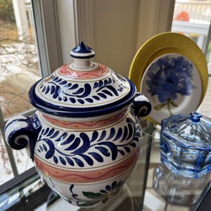 May include: A decorative ceramic jar with a lid, adorned with a blue and white floral pattern and pink details. The jar has two handles and is placed on a glass surface. A yellow plate and a blue floral plate are visible.