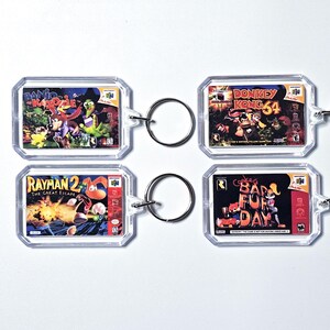 Peut inclure: Quatre porte-clés rectangulaires en acrylique transparent présentant des miniatures de jaquettes de jeux Nintendo 64 : Banjo-Kazooie, Donkey Kong 64, Rayman 2: The Great Escape et Conker's Bad Fur Day, chacun fixé à un anneau en métal argenté.
