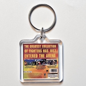 May include: A square acrylic keychain featuring a miniature replica of a video game case back cover. The design includes yellow text reading THE GREATEST COLLECTION OF FIGHTERS HAS JUST ENTERED THE ARENA, game screenshots, a barcode, and a Teen rating.
