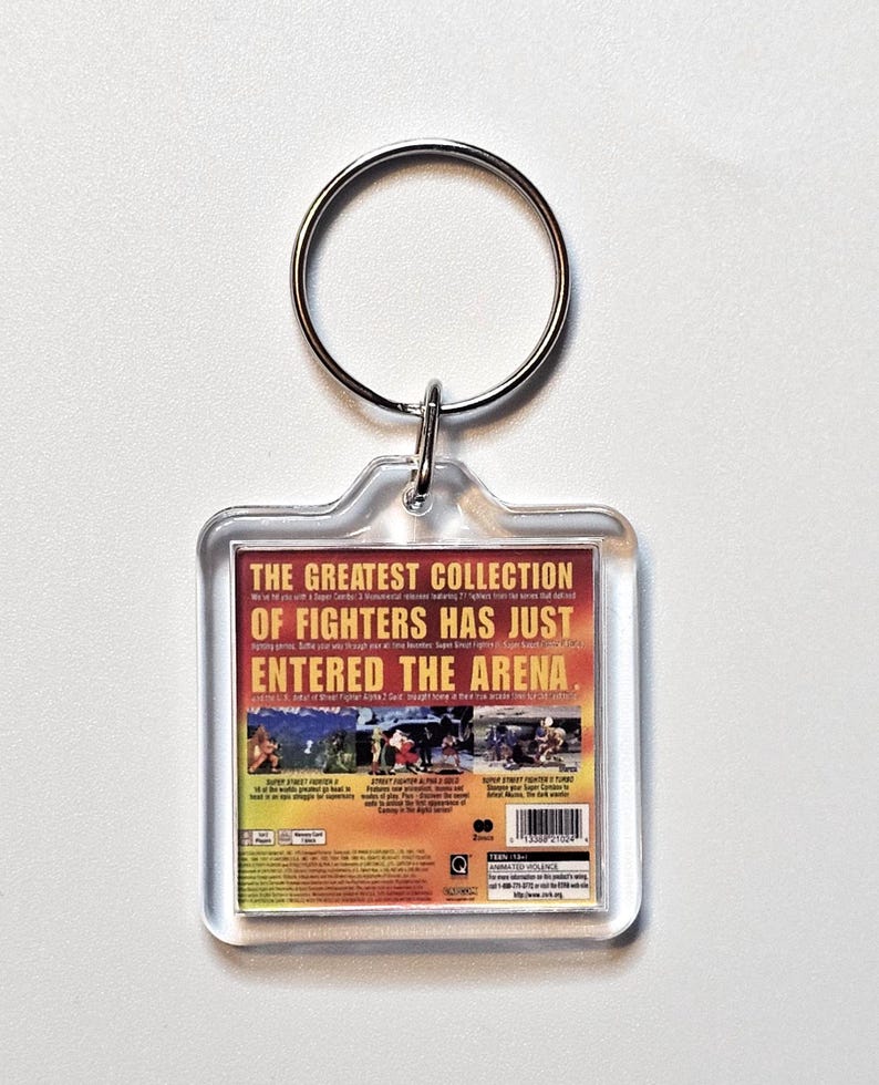 May include: A square acrylic keychain featuring a miniature replica of a video game case back cover. The design includes yellow text reading THE GREATEST COLLECTION OF FIGHTERS HAS JUST ENTERED THE ARENA, game screenshots, a barcode, and a Teen rating.