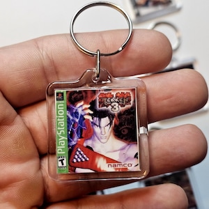 May include: A square clear acrylic keychain featuring a miniature replica of the Tekken 3 video game cover art for PlayStation. The graphic displays the game title, the Namco logo, and a Teen ESRB rating. A silver metal split ring is attached to the top.