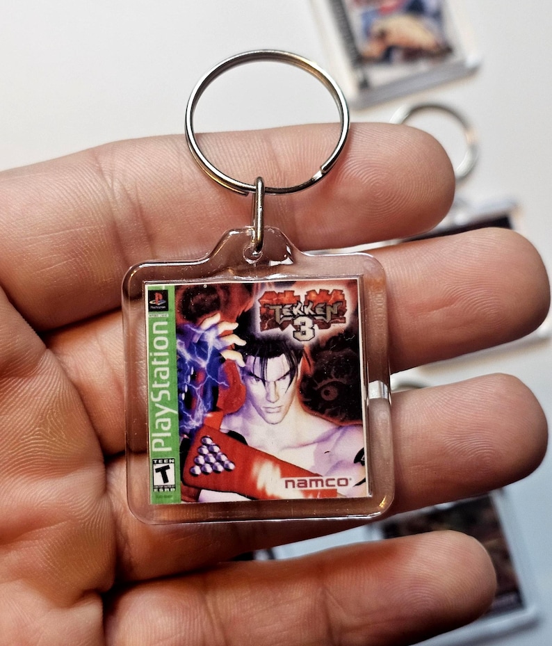 May include: A square clear acrylic keychain featuring a miniature replica of the Tekken 3 video game cover art for PlayStation. The graphic displays the game title, the Namco logo, and a Teen ESRB rating. A silver metal split ring is attached to the top.