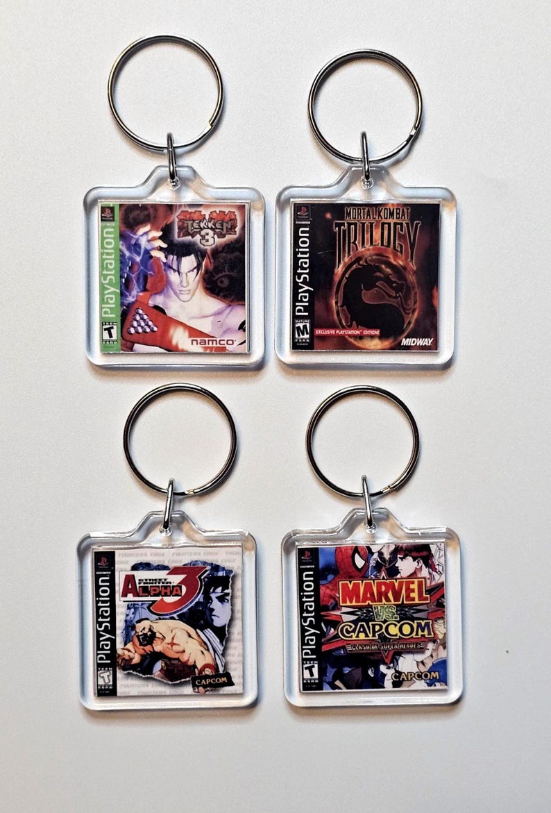 May include: Four square acrylic keychains featuring miniature PlayStation game cover art for Tekken 3, Mortal Kombat Trilogy, Street Fighter Alpha 3, and Marvel vs. Capcom. Each keychain includes a silver metal split ring for attachment.