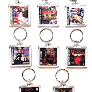 May include: Eight square clear acrylic keychains featuring miniature PlayStation game cover art, including Tekken 3, Marvel vs. Capcom, Street Fighter Alpha 3, Tekken, Mortal Kombat Trilogy, Bloody Roar, Tekken 2, and Soul Blade, each with a metal ring.