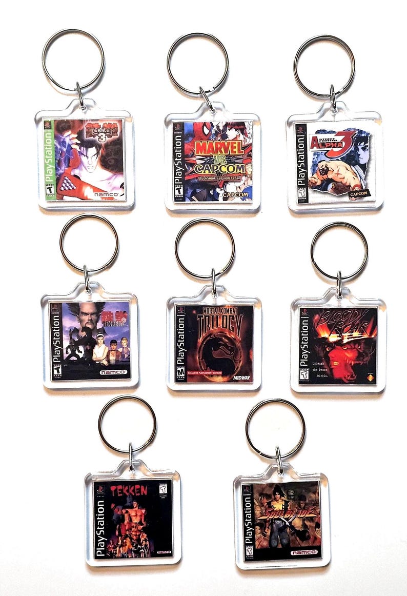 May include: Eight square clear acrylic keychains featuring miniature PlayStation game cover art, including Tekken 3, Marvel vs. Capcom, Street Fighter Alpha 3, Tekken, Mortal Kombat Trilogy, Bloody Roar, Tekken 2, and Soul Blade, each with a metal ring.