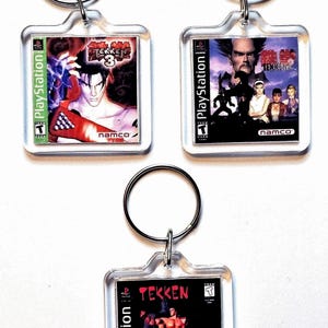 May include: Three square clear acrylic keychains featuring miniature PlayStation game cover art for Tekken 3, Tekken 2, and the original Tekken. Each keychain includes a silver metal split ring attached to the top for securing keys.