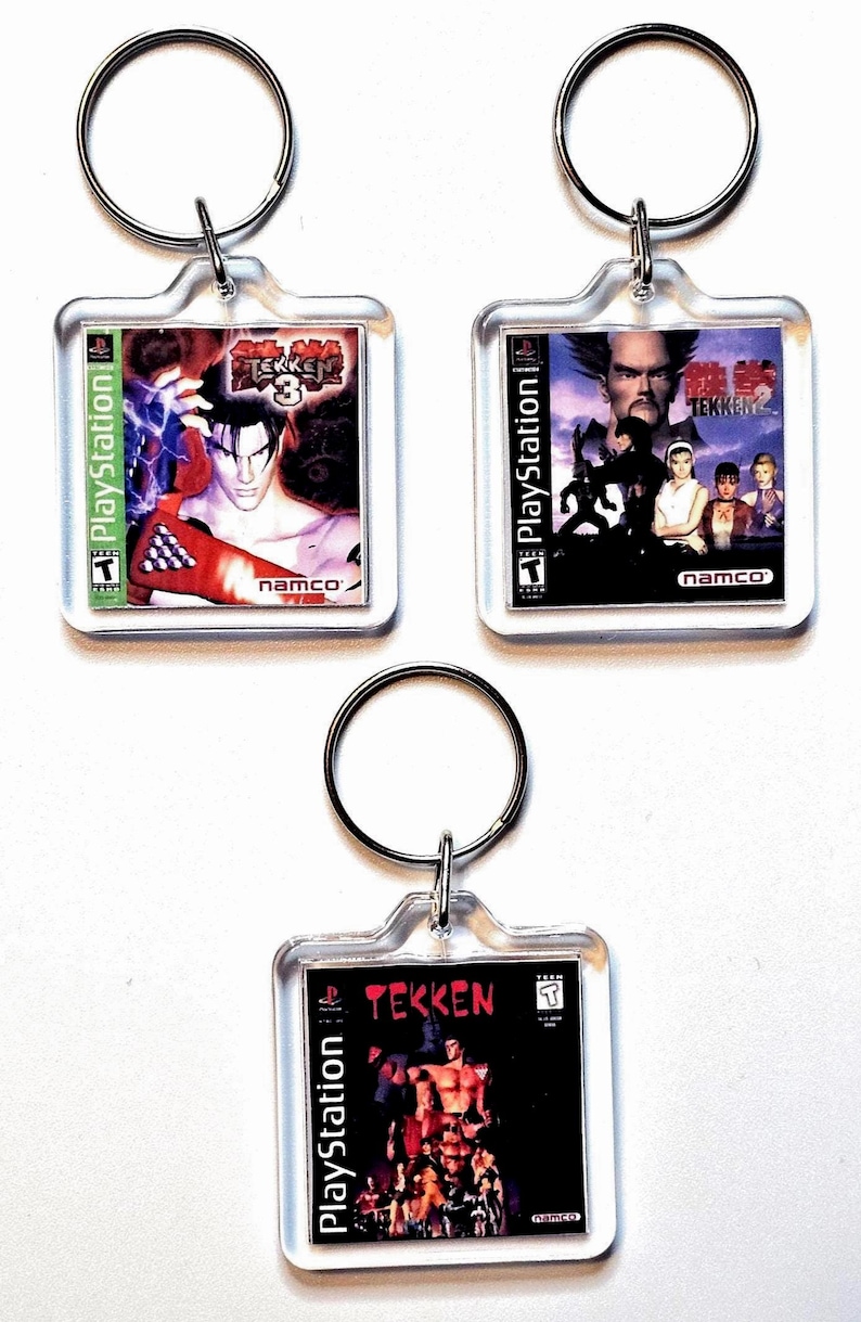May include: Three square clear acrylic keychains featuring miniature PlayStation game cover art for Tekken 3, Tekken 2, and the original Tekken. Each keychain includes a silver metal split ring attached to the top for securing keys.