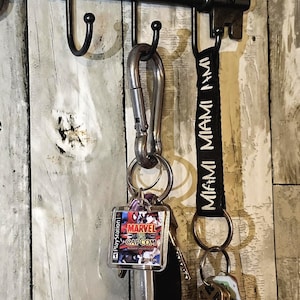 May include: A metal key rack with a black finish holding two keychains. One keychain features a silver carabiner with a square Marvel vs. Capcom PlayStation game case charm. The other is a black fabric strap with the word MIAMI printed in white.