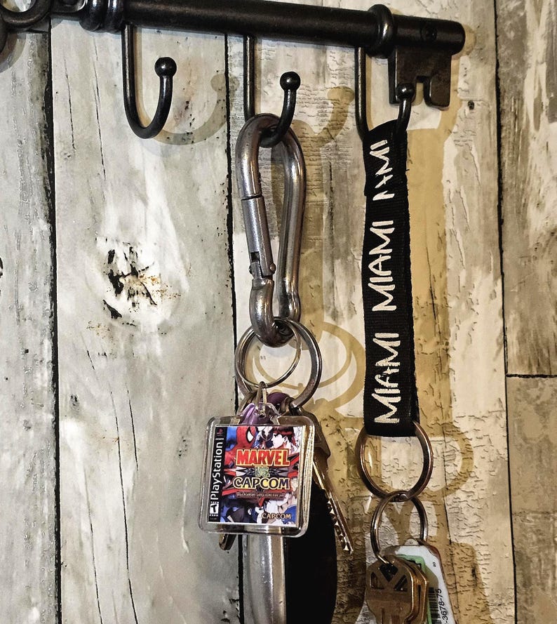 May include: A metal key rack with a black finish holding two keychains. One keychain features a silver carabiner with a square Marvel vs. Capcom PlayStation game case charm. The other is a black fabric strap with the word MIAMI printed in white.