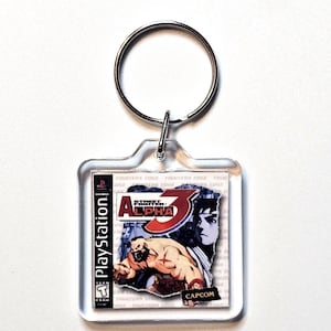 May include: A square clear acrylic keychain featuring a miniature replica of the Street Fighter Alpha 3 PlayStation game cover art. The design includes the game title, the Capcom logo, and a graphic illustration of Zangief. A silver metal ring is attached.
