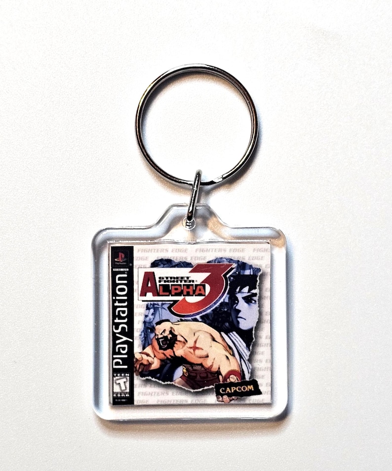 May include: A square clear acrylic keychain featuring a miniature replica of the Street Fighter Alpha 3 PlayStation game cover art. The design includes the game title, the Capcom logo, and a graphic illustration of Zangief. A silver metal ring is attached.