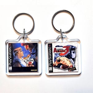 May include: Two square clear acrylic keychains featuring miniature PlayStation game cover art. The left keychain displays Street Fighter Alpha 2, and the right keychain displays Street Fighter Alpha 3, both with metal rings for attachment.