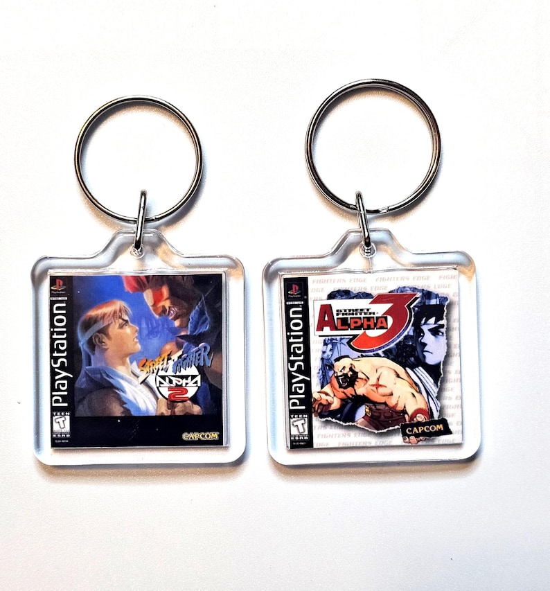 May include: Two square clear acrylic keychains featuring miniature PlayStation game cover art. The left keychain displays Street Fighter Alpha 2, and the right keychain displays Street Fighter Alpha 3, both with metal rings for attachment.