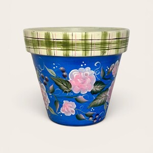 May include: A hand-painted terracotta flower pot featuring a vibrant blue base decorated with pink roses, green leaves, and dark berries. The rim is painted with a green and white plaid pattern consisting of thin horizontal and vertical lines.
