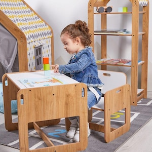 Può includere: Un set tavolo e sedia per bambini in legno chiaro con piano e seduta bianchi. Il set è abbinato a una libreria e a una tenda gioco. Il bambino gioca con blocchi colorati sul tavolo.