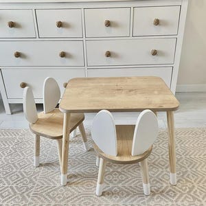 Puede incluir: Un juego de mesa y dos sillas para niños de madera clara. Las sillas tienen respaldos blancos en forma de oreja. Las patas de la mesa y las sillas son de madera clara con puntas blancas. El conjunto está sobre una alfombra estampada.