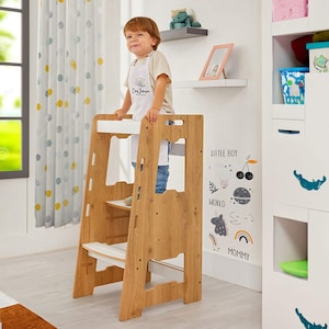 Puede incluir: Un taburete de madera para niños pequeños con detalles en blanco. El taburete tiene tres niveles y una barra de seguridad. El niño lleva un delantal blanco con el nombre "Cay Johnson". El fondo incluye un armario blanco y una calcomanía de pared.