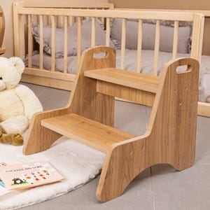 Può includere: Uno sgabello in legno a due gradini con finitura naturale. Lo sgabello ha una maniglia tagliata nella parte superiore di ogni lato. I gradini sono rettangolari e le gambe hanno un design curvo. Un libro per bambini e un orsacchiotto sono sullo sfondo.