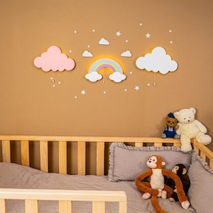 Peut inclure: Décoration murale de chambre d'enfant avec un arc-en-ciel et des nuages lumineux, avec des étoiles. L'arc-en-ciel est aux couleurs pastel. Un lit bébé en bois est au premier plan, avec des peluches dont un ours en peluche et des singes.