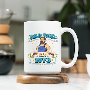Op de afbeelding: Witte keramische mok met de tekst "DAD BOD: LIMITED EDITION SINCE 1973" en een cartoon illustratie van een man in een blauwe overall. De mok staat op een houten onderzetter.