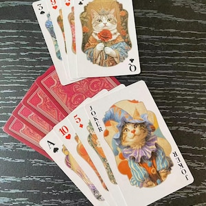 Puede incluir: Una baraja de cartas con ilustraciones caprichosas de gatos. Las cartas incluyen una Reina sosteniendo una rosa y un Joker con un sombrero de bufón. Los dorsos de las cartas son rojos con un diseño floral. Las cartas están dispuestas sobre una superficie oscura.
