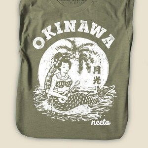 May include: Olive green t-shirt featuring a white graphic of a mermaid playing a ukulele in front of a full moon and palm trees. The word "OKINAWA" is printed above the graphic. The brand name "Moshi Moshi" is printed on the collar.
