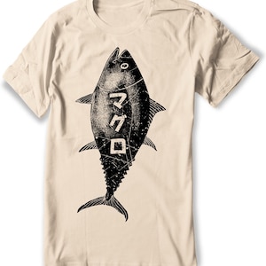 Puede incluir: Una camiseta beige con una ilustraci&oacute;n en blanco y negro de un at&uacute;n. El pez tiene la palabra japonesa "マグロ" (maguro) escrita en &eacute;l, que significa at&uacute;n.
