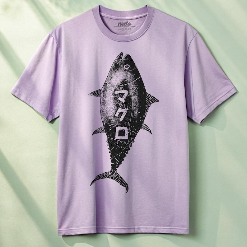 Puede incluir: Una camiseta de color lavanda con un gr&aacute;fico en blanco y negro de un at&uacute;n con la palabra japonesa "マグロ" (maguro, que significa at&uacute;n) impresa en ella.