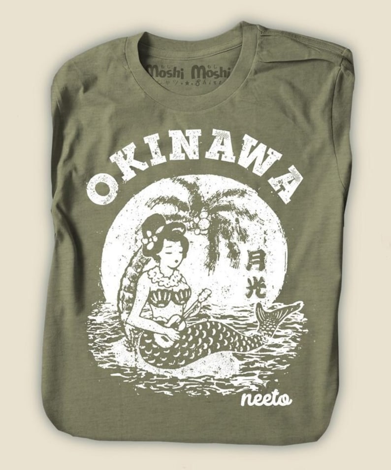 May include: Olive green t-shirt featuring a white graphic of a mermaid playing a ukulele in front of a full moon and palm trees. The word "OKINAWA" is printed above the graphic. The brand name "Moshi Moshi" is printed on the collar.